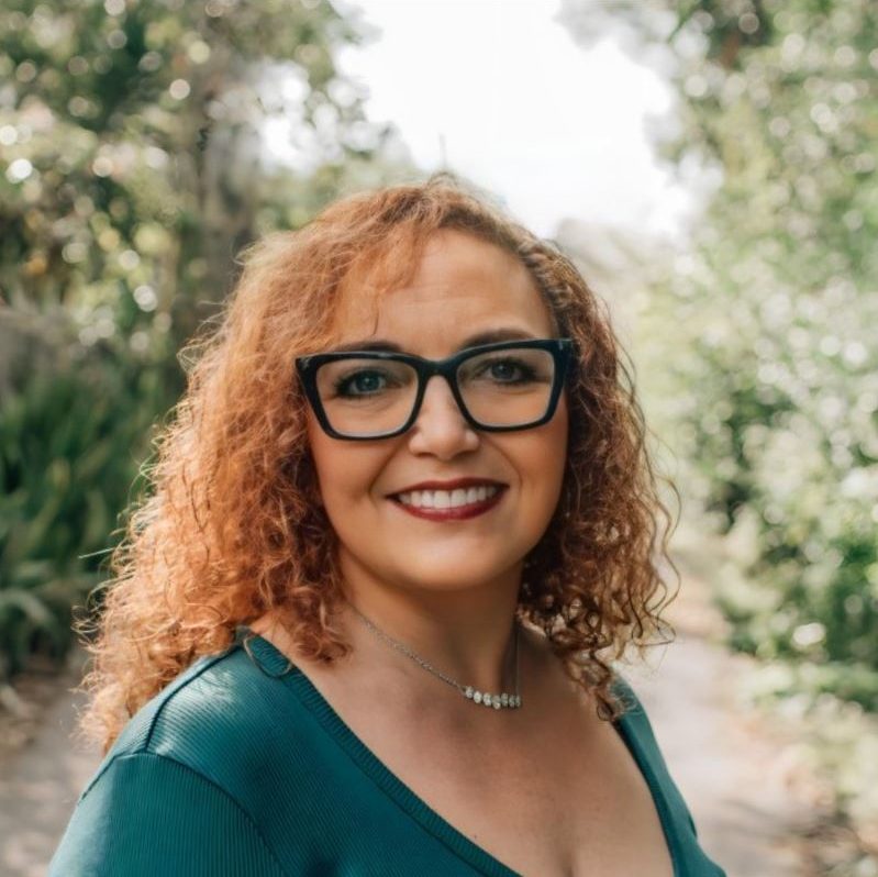 A woman with red hair and glasses stands amidst lush greenery in a serene forest setting.