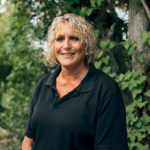 A woman in a black shirt stands before trees, symbolizing her journey to improve access to compassionate caregiving services.