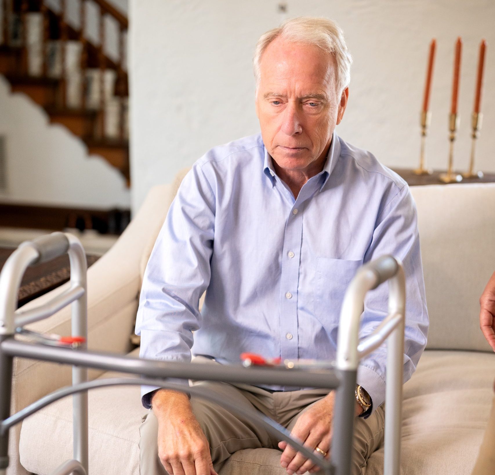 An older man sitting on a couch, using a walker for support, reflecting post-surgery recovery at home.