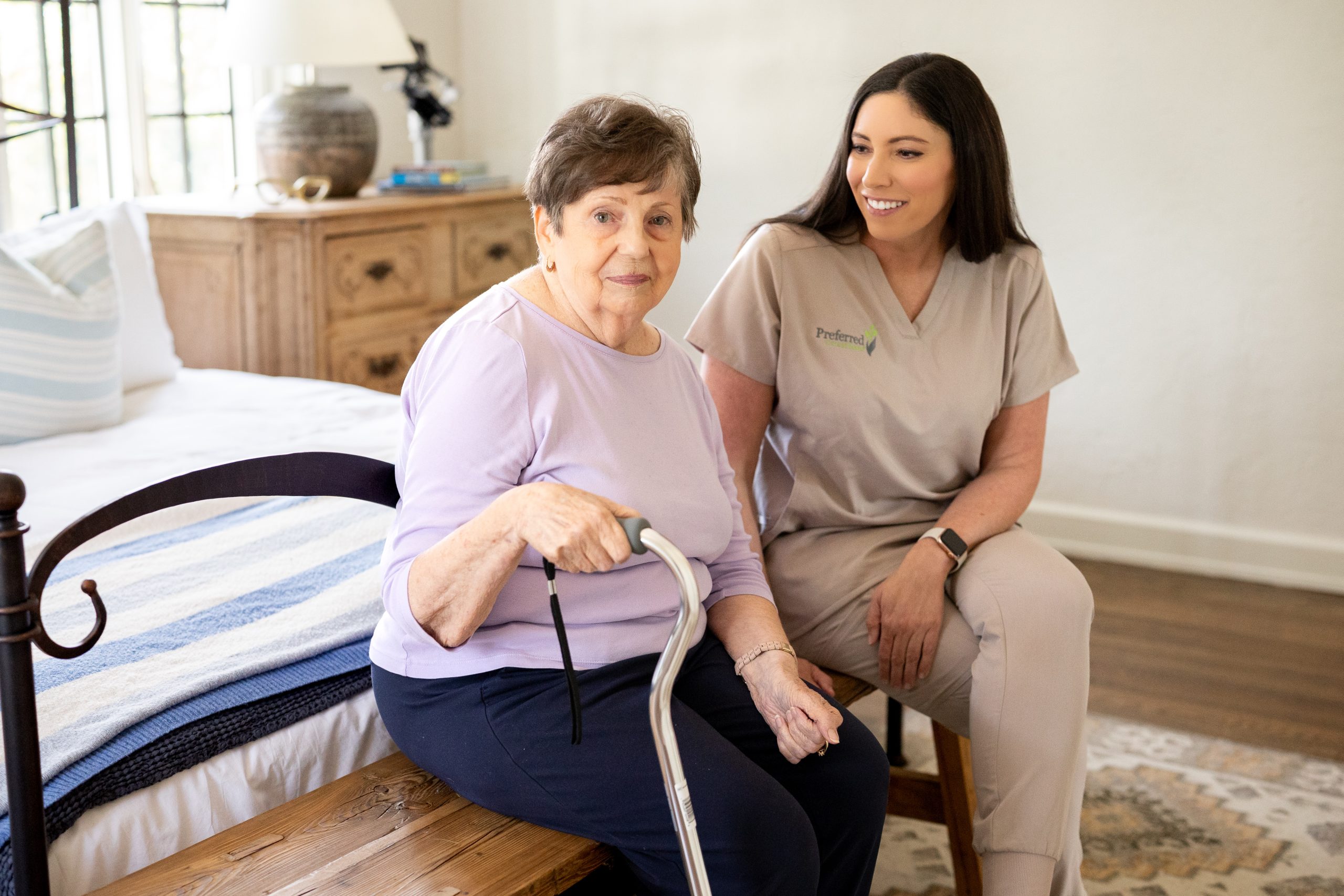 A woman with a cane stands beside another woman sitting on a bed, symbolizing support and companionship in home care.