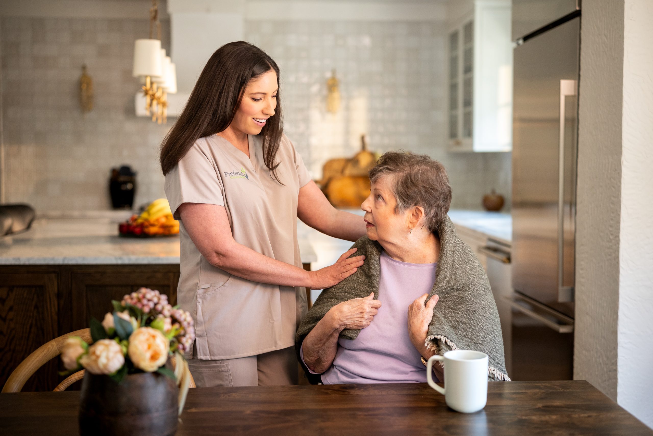 A woman in a nursing uniform supports an older woman in a kitchen, exemplifying compassionate in-home care for seniors.
