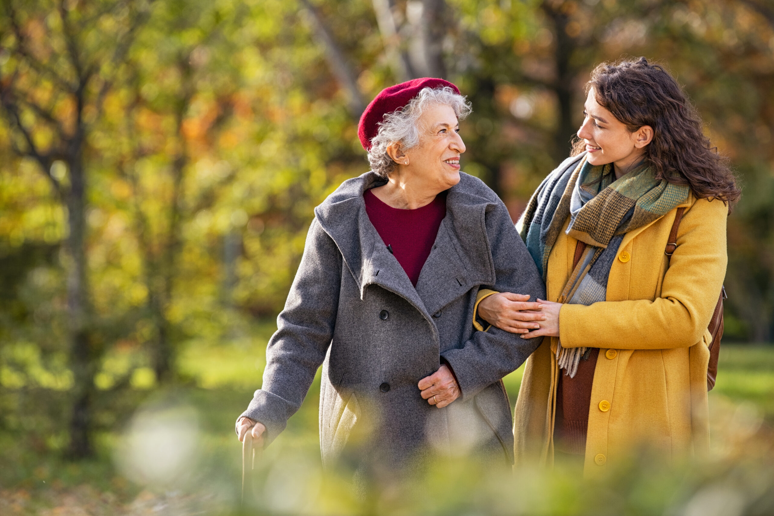 Smiling senior woman in a red beret and gray coat walking arm-in-arm with her granddaughter through a park during autumn — a warm afternoon walk together