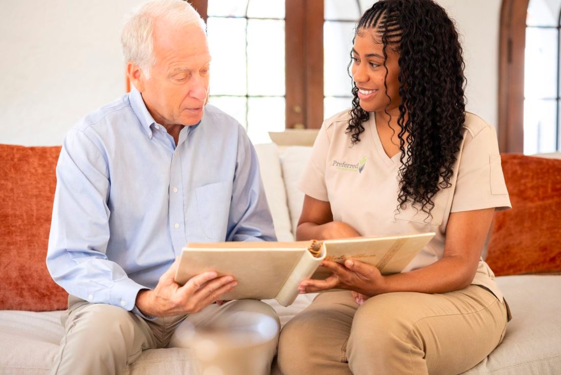 A woman and an older man sit on a couch, engaged in reading a book together, highlighting Alzheimer's care activities.