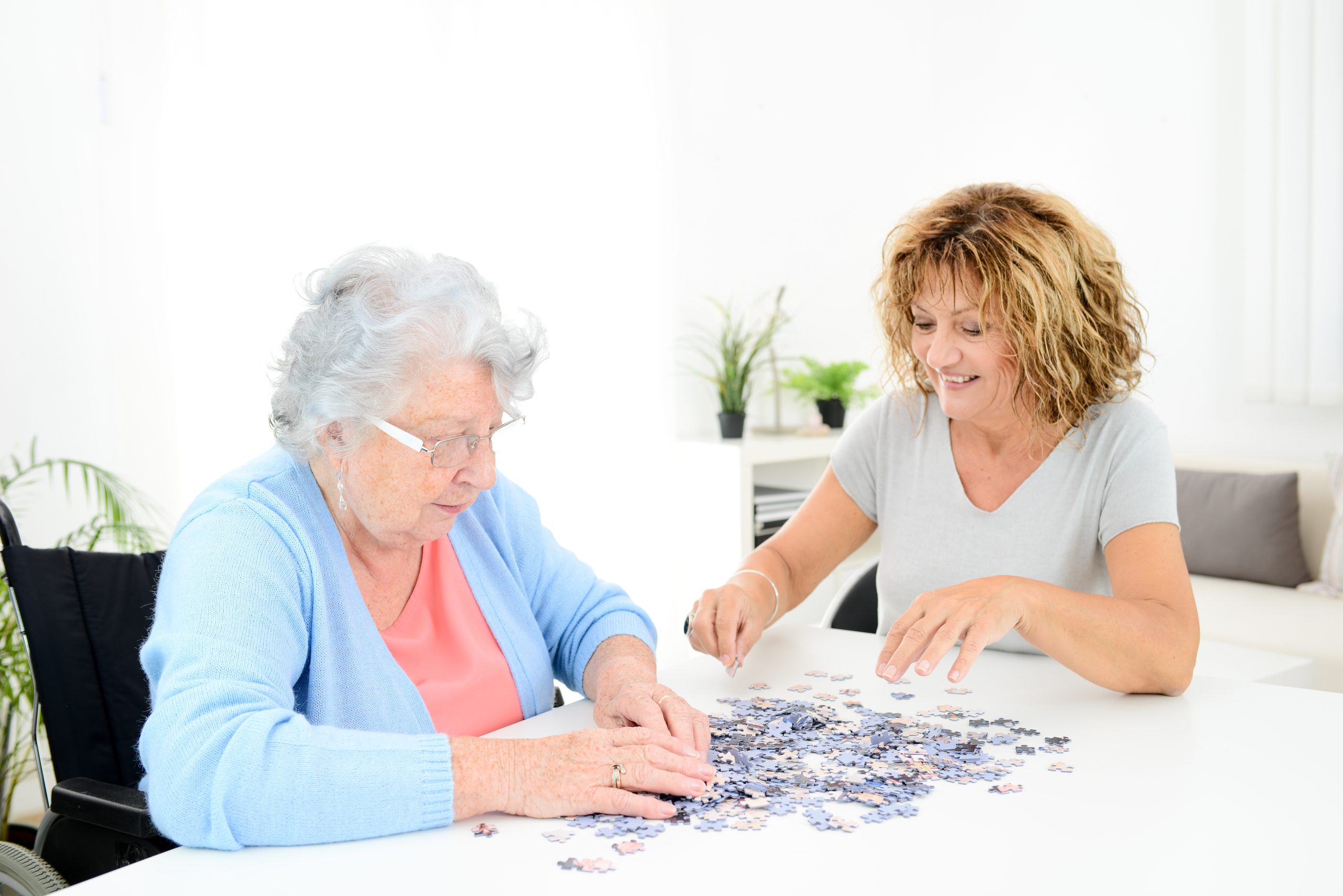 Two senior women smiling and working on a jigsaw puzzle together in a sunlit kitchen, capturing a calm, grounded daily routine and warm companionship