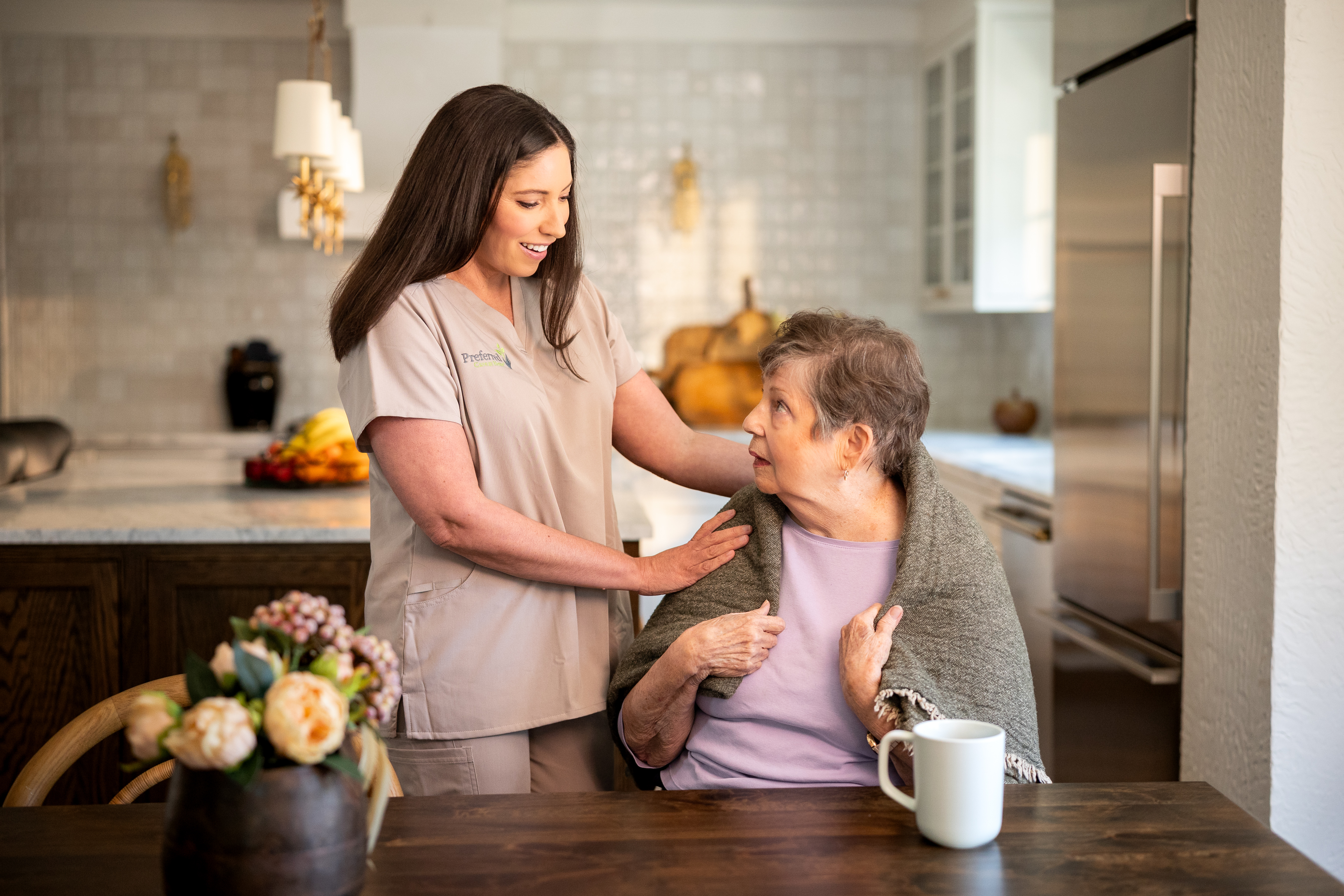 A caregiver in a nursing uniform supports an older woman in a kitchen, the kind of compassionate, dependable in-home care families look for.
