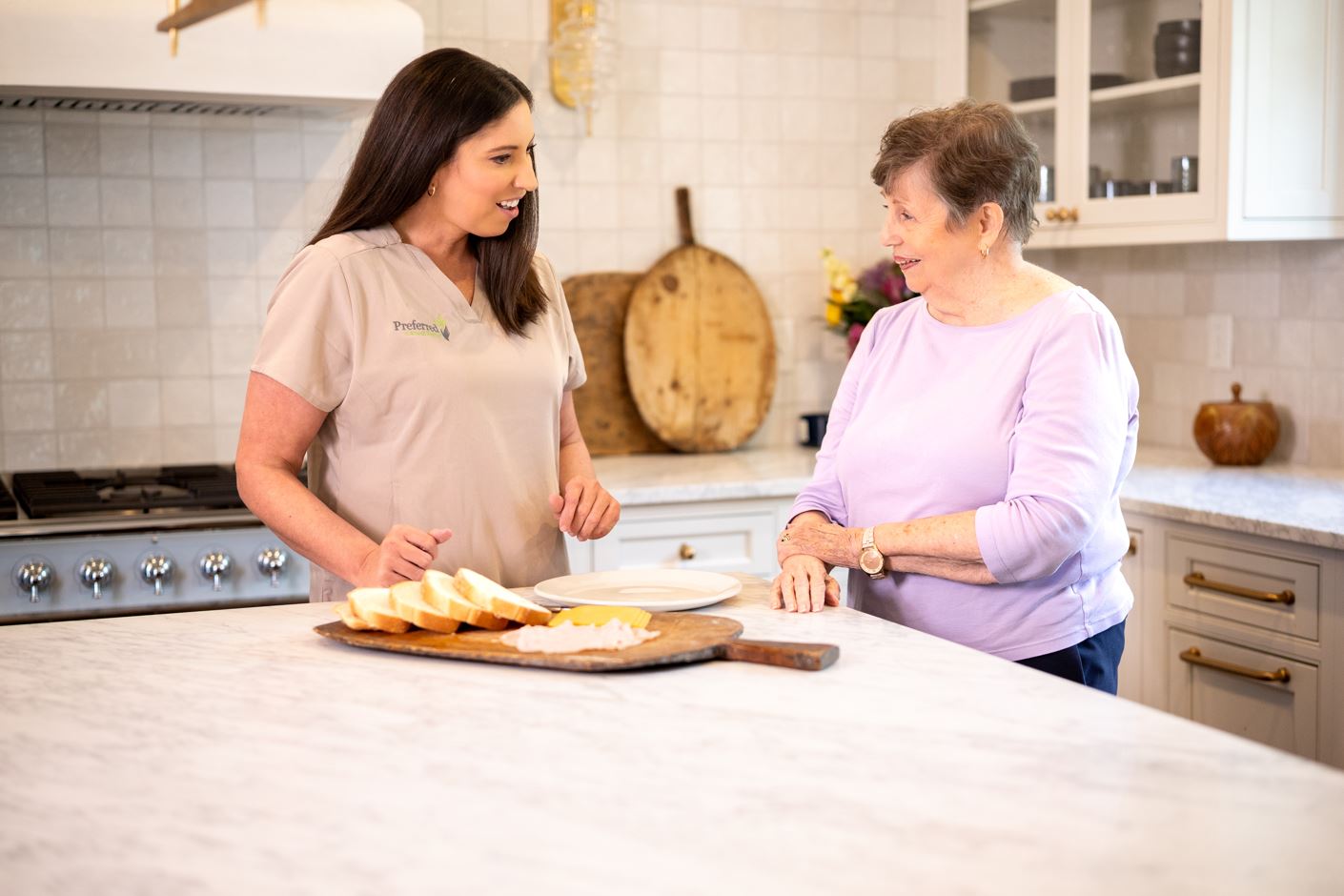 A woman prepares food in a kitchen for an elderly woman seated nearby, showcasing care and assistance in meal preparation.