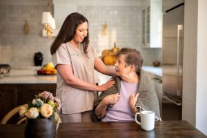 A woman in nursing attire helps an older woman in a kitchen, showcasing compassion and assistance in daily tasks.