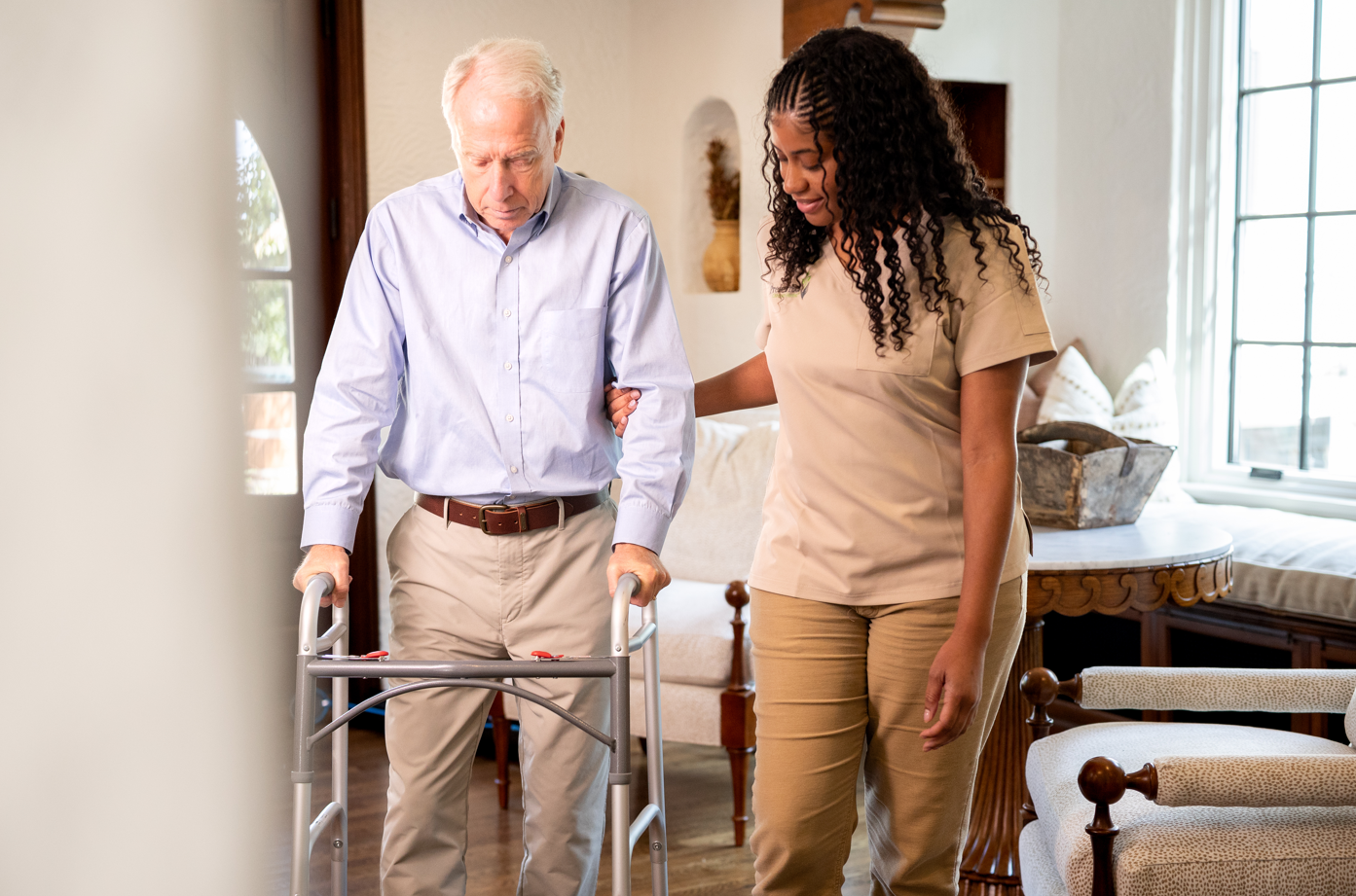 A woman assists an elderly man using a walker in a home setting, highlighting post-surgery care.