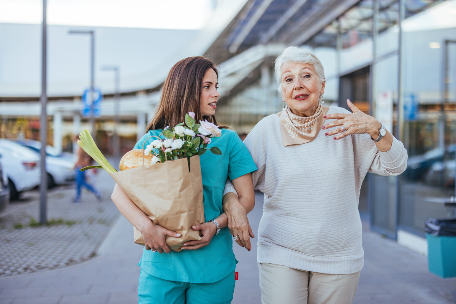 A young caregiver and an elderly woman with white hair walk together after shopping. The caregiver carries a bag of groceries and flowers.