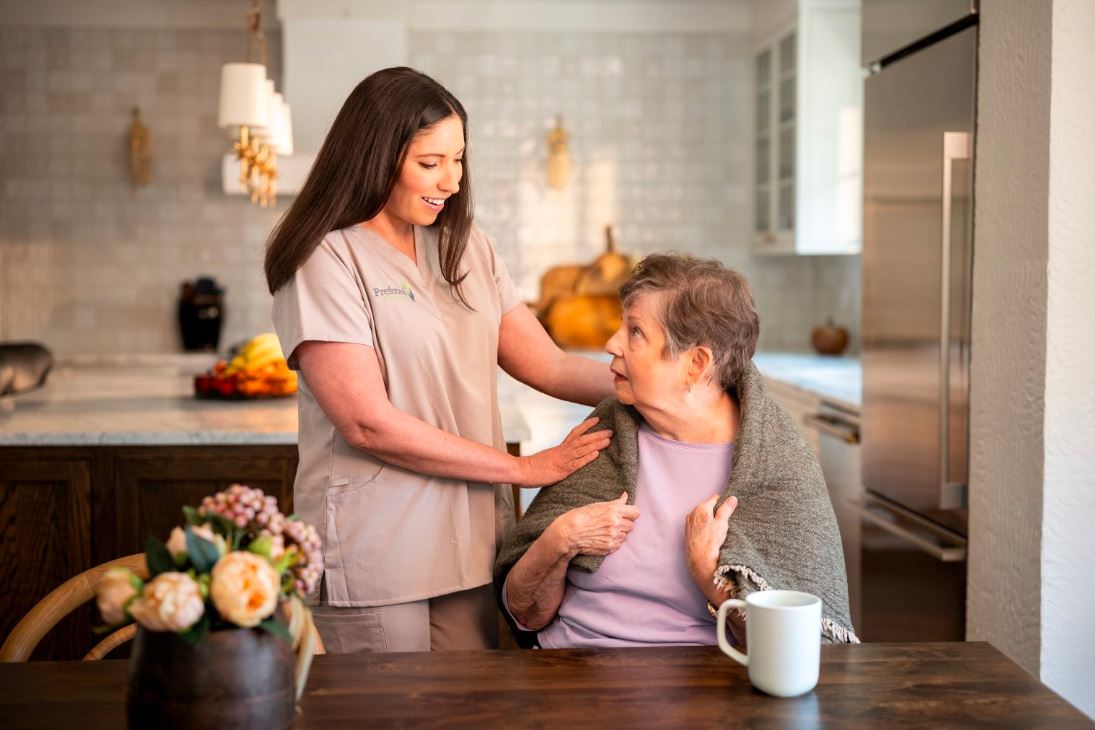 A woman in a nursing uniform supports an older woman in a kitchen, exemplifying compassionate in-home care for seniors.