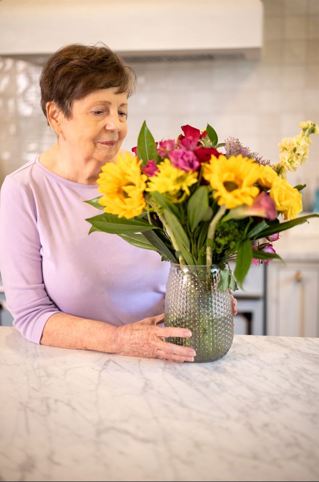 An older woman smiles while holding a colorful vase filled with fresh flowers.