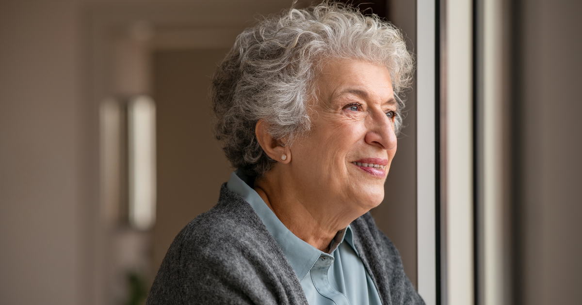 elderly woman looking out the window smiling while wearing a blue shirt.